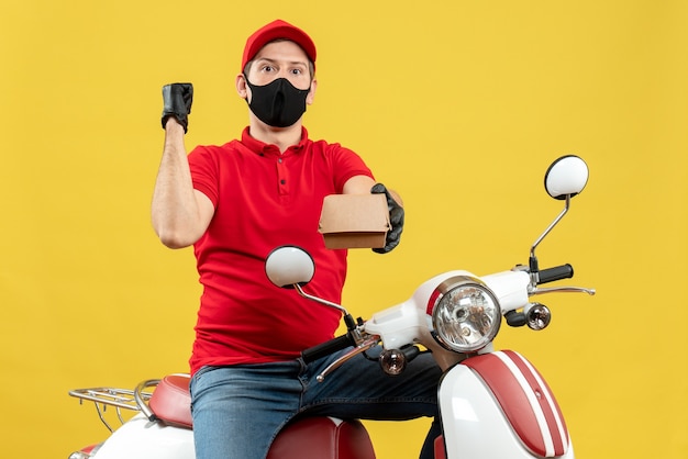 Vista superior del hombre de mensajería vestido con blusa roja y guantes de sombrero en máscara médica sentado en scooter sosteniendo orden apuntando hacia atrás