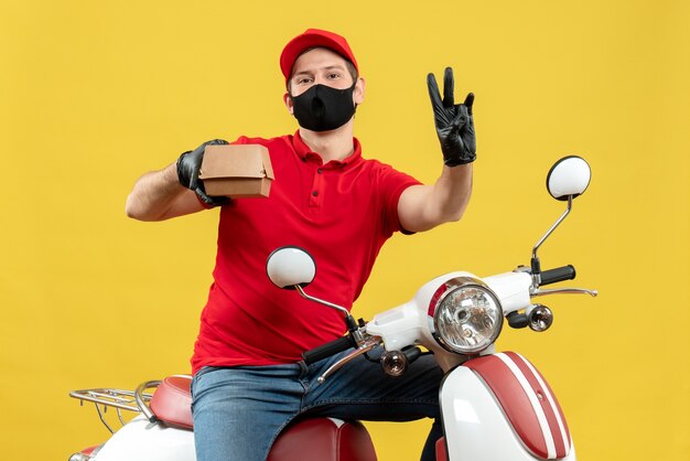 Vista superior del hombre de mensajería vestido con blusa roja y guantes de sombrero en máscara médica sentado en scooter sosteniendo orden mostrando tres