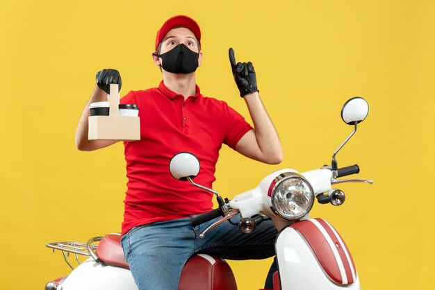 Vista superior del hombre de mensajería vestido con blusa roja y guantes de sombrero en máscara médica sentado en scooter sosteniendo órdenes apuntando hacia arriba