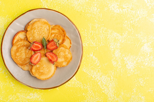 Vista superior horneados deliciosos panqueques dentro de la placa marrón con fresas en rodajas en la pared amarilla postre dulce baya de alimentos para panqueques