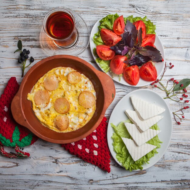 Vista superior huevos fritos con salchichas con tomate picado y queso y un vaso de té en plato de arcilla