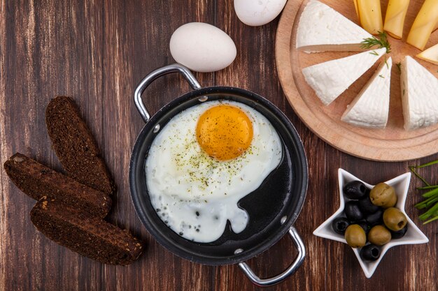 Foto gratuita vista superior de huevos fritos en una sartén con aceitunas y cebollas verdes, pan negro y queso feta sobre un fondo de madera