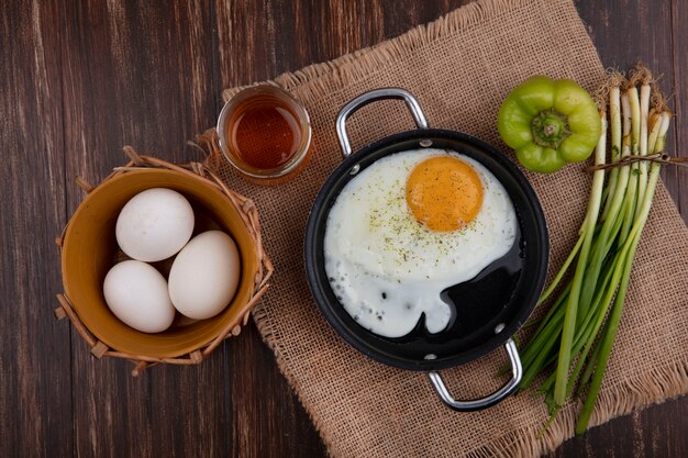 Vista superior de huevos fritos en una sartén con miel cebollas verdes pimientos y huevos de gallina en una canasta sobre un fondo de madera