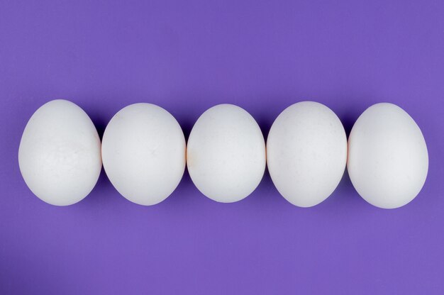 Vista superior de huevos de gallina blancos frescos y saludables dispuestos en una línea sobre un fondo violeta