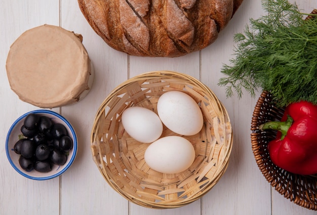 Vista superior de huevos de gallina en canasta con aceitunas yogur hogaza de pan con eneldo sobre fondo blanco.