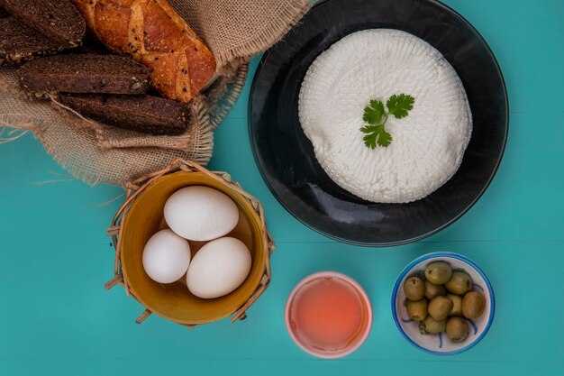 Vista superior de huevos de gallina en una canasta con queso, aceitunas y pan negro sobre un fondo turquesa