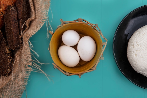 Vista superior de huevos de gallina en una canasta con queso y pan negro sobre un fondo turquesa