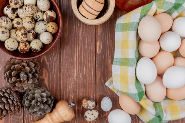 Vista superior de huevos de gallina sobre un mantel marcado y huevos de codorniz en un recipiente con piñas aisladas sobre un fondo de madera