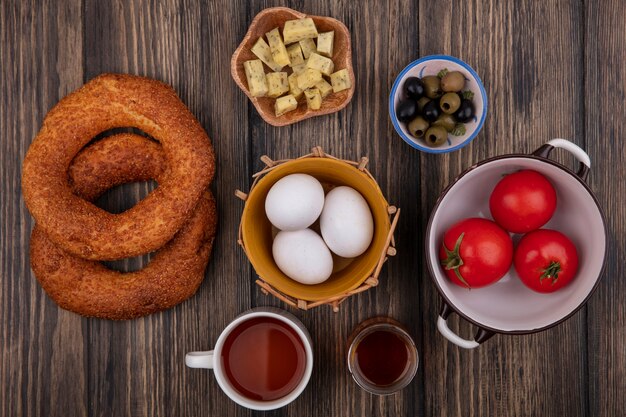 Vista superior de huevos orgánicos en un balde con queso en un cuenco de madera con tomates en un cuenco y con una taza de té sobre un fondo de madera