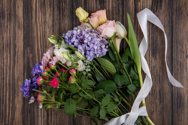 Vista superior de increíbles flores como margaritas de rosas lilas con hojas con cinta blanca sobre un fondo de madera