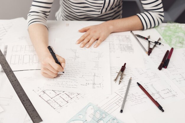 Vista superior del ingeniero independiente joven y guapo con ropa formal a rayas que trabaja en una cómoda mesa bif, tomando notas cerca de planos para arreglarlos más tarde.