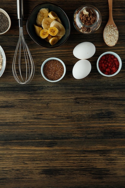 Vista superior de los ingredientes del desayuno como grosella roja de sésamo de huevo de nuez de plátano con batidor sobre fondo de madera con espacio de copia