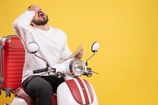 Vista superior del joven agotado sentado en la motocicleta con la maleta en él sosteniendo el mapa sobre fondo amarillo aislado