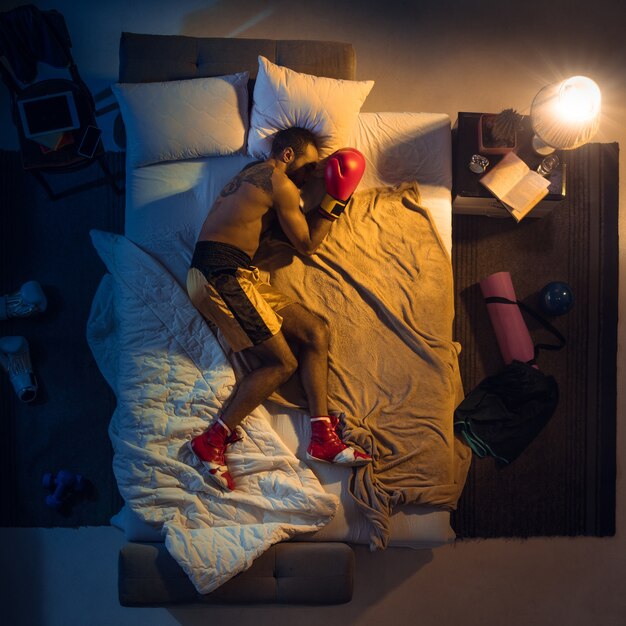 Vista superior del joven boxeador profesional, luchador durmiendo en su habitación en ropa deportiva con guantes