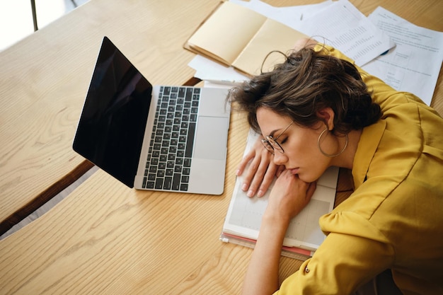 Vista superior de una joven cansada soñadora que se queda dormida en el escritorio con una computadora portátil y documentos debajo de la cabeza en el lugar de trabajo