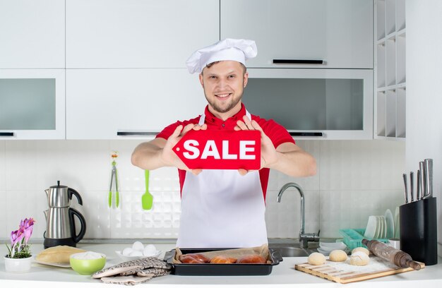 Vista superior del joven chef masculino sonriente mostrando cartel de venta en la cocina blanca