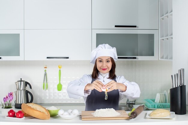 Vista superior de la joven chef mujer sonriente en uniforme de pie detrás de la mesa rompiendo el huevo en la comida en la cocina blanca