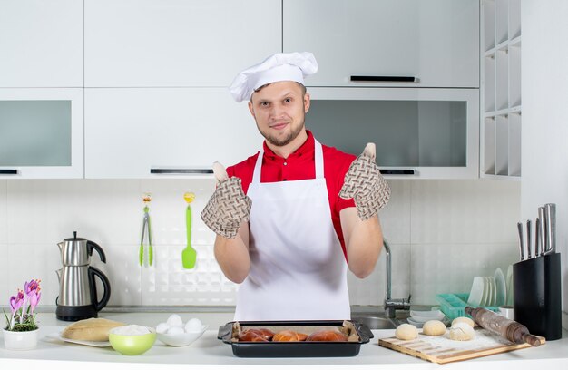 Vista superior del joven chef con soporte de pie detrás de la mesa con pasteles rallador de huevos y haciendo un gesto de ok en la cocina blanca