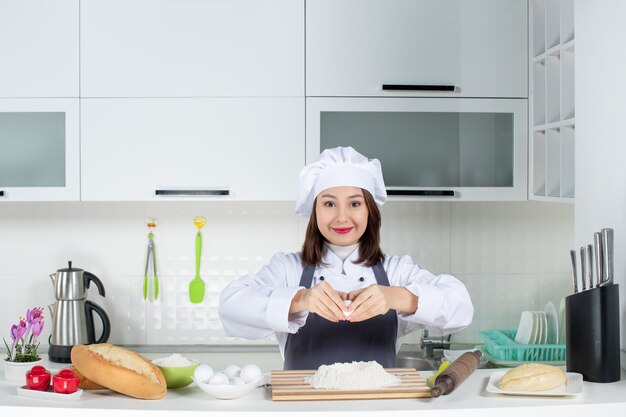 Vista superior de la joven chef en uniforme de pie detrás de la mesa rompiendo el huevo en la comida en la cocina blanca