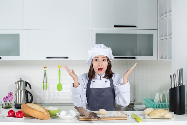 Vista superior de la joven cocinera de comis sorprendida en uniforme de pie detrás de la mesa manchando su rostro con harina en la cocina blanca