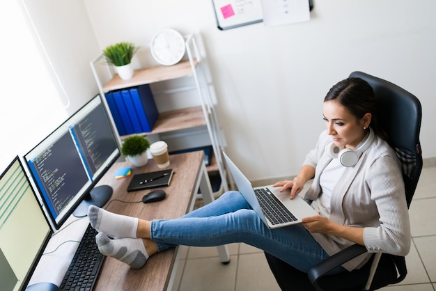 Vista superior de una joven descalza descansando los pies en el escritorio de su oficina mientras trabaja en la codificación de una aplicación de software desde casa