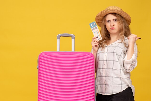 Vista superior de una joven emocional con sombrero que muestra el boleto y de pie cerca de su bolso rosa apuntando hacia atrás