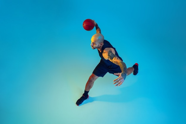 Vista superior. Joven jugador de baloncesto del equipo vistiendo ropa deportiva, practicando en acción, movimiento sobre fondo azul en luz de neón. Concepto de deporte, movimiento, energía y estilo de vida dinámico y saludable.