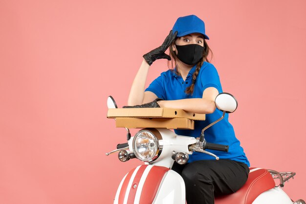 Vista superior del joven mensajero femenino orgulloso con máscara médica y guantes sentado en scooter entregando pedidos en melocotón pastel