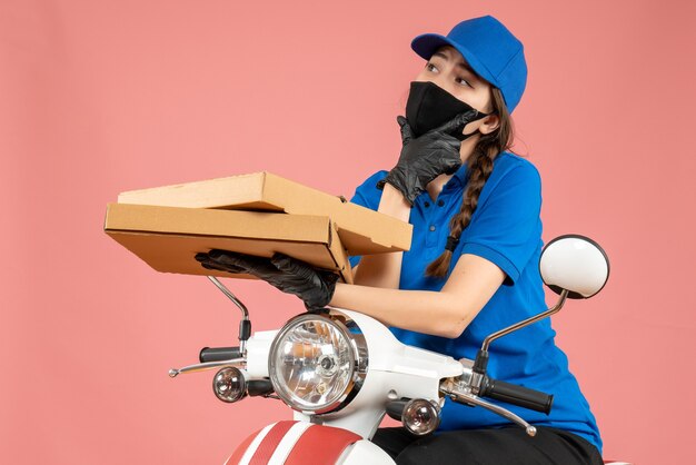 Vista superior del joven mensajero femenino reflexivo con máscara médica y guantes sosteniendo cajas en melocotón pastel