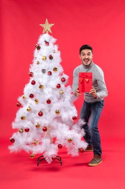 Vista superior del joven parado cerca del árbol de año nuevo blanco decorado y sosteniendo sus regalos y mostrándoselo a sus amigos en rojo