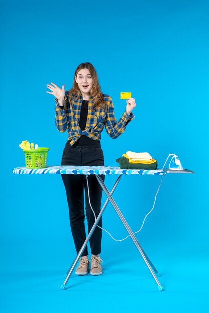 Vista superior de la joven que muestra la tarjeta bancaria sintiéndose conmocionada y de pie detrás de la tabla de planchar en el lavadero