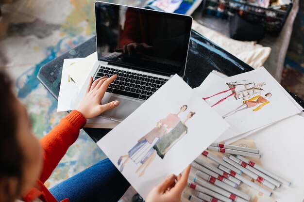 Vista superior de una joven sentada en la mesa trabajando en una laptop con ilustraciones de moda cerca de un taller moderno