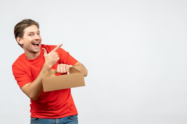 Vista superior del joven sonriente y emocional en blusa roja con caja apuntando algo en el lado izquierdo en la pared blanca