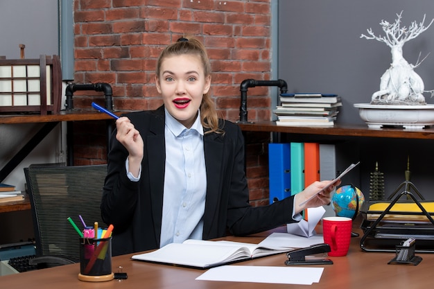 Foto gratuita vista superior de la joven sorprendida sentada en una mesa y sosteniendo un documento con bolígrafo de color azul en la oficina