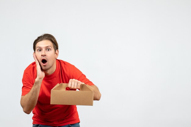 Vista superior del joven sorprendido y emocional en blusa roja con caja sobre fondo blanco.