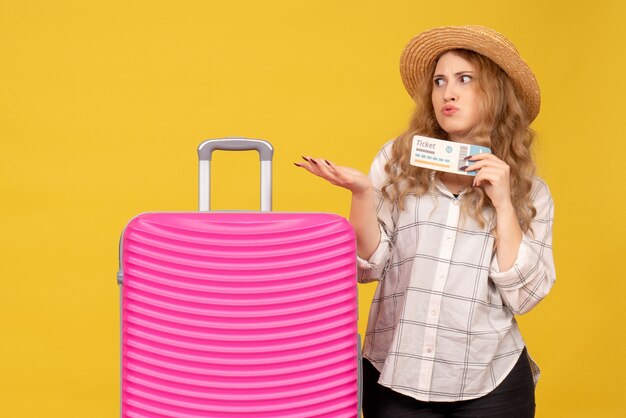 Vista superior de la jovencita preguntándose con sombrero sosteniendo boleto y de pie cerca de su bolso rosa