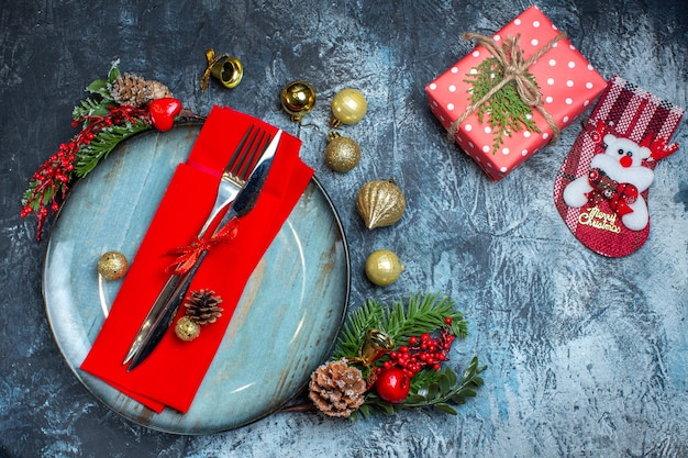 Vista superior del juego de cubiertos con cinta roja en una servilleta decorativa en un plato azul y accesorios navideños, caja de regalo de cono de conífera y calcetín navideño sobre fondo oscuro