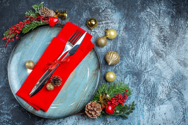 Vista superior del juego de cubiertos con cinta roja en una servilleta decorativa sobre un plato azul y accesorios navideños en el lado derecho sobre fondo oscuro