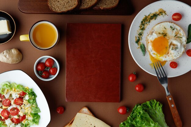 Vista superior del juego de desayuno con huevo frito con tomate y eneldo y ensalada de verduras con lechuga de huevo de tomate con rebanadas de pan de jugo de naranja tenedor y tabla de cortar en el centro