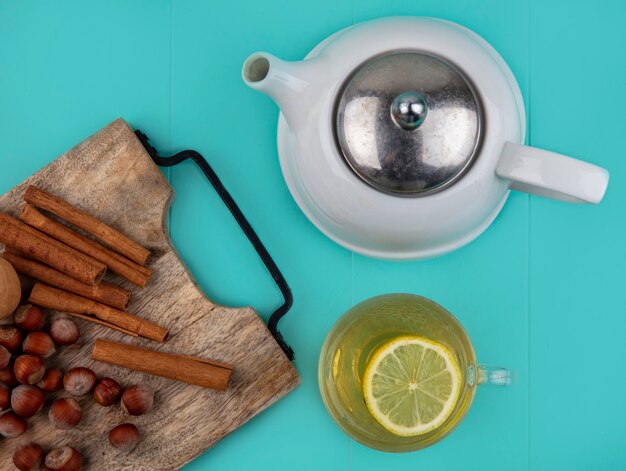 Vista superior de jugo de limón con una rodaja de limón en un vaso y nueces canela nuez sobre tabla de cortar con tetera sobre fondo azul.