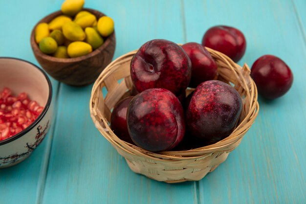 Vista superior de jugosos pluots en un balde con semillas de granada en un recipiente con kinkans en un recipiente de madera sobre un fondo de madera azul