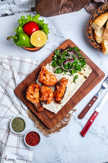 Vista superior de kebab de pollo con hierbas y cebolla roja en lavash sobre tabla de madera servido con verduras frescas y especias en la mesa