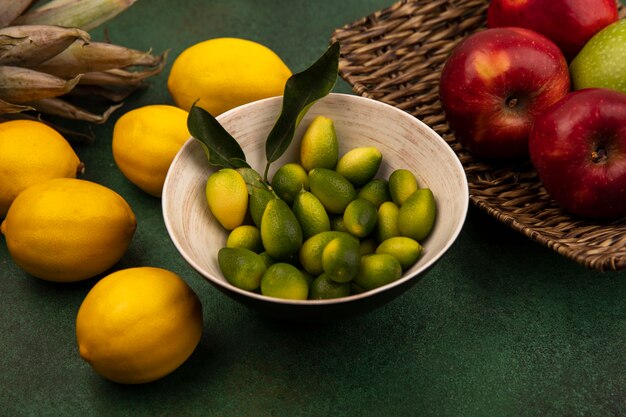 Vista superior de kinkans aromáticos en un recipiente con manzanas rojas frescas en una bandeja de mimbre con limones aislado en una pared verde