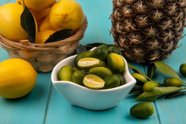 Vista superior de kinkans frescos en un recipiente con limones en un cubo con piña aislado sobre un fondo de madera azul