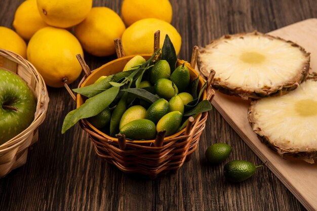 Vista superior de kinkans verdes con hojas en un balde con piñas en una tabla de cocina de madera con manzanas en balde con limones aislado en una superficie de madera