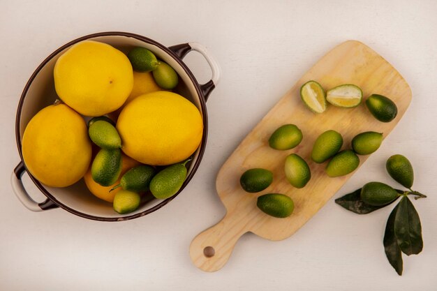 Vista superior de kinkans verdes en un recipiente con limones con medio kinkans aislado en una tabla de cocina de madera sobre una pared blanca