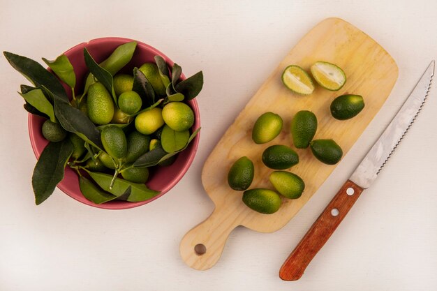 Vista superior de kinkans verdes en un recipiente con medio kinkans aislado en una tabla de cocina de madera con un cuchillo en una pared blanca