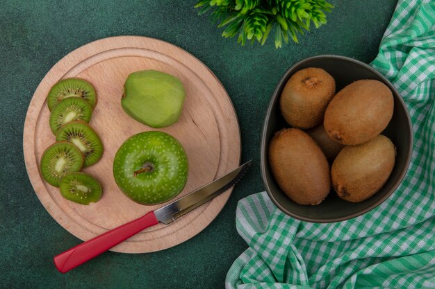 Vista superior de kiwi en un recipiente con una manzana verde y un cuchillo en un soporte con una toalla a cuadros verde sobre un fondo verde