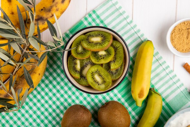 Vista superior de kiwi en rodajas en un tazón, frutas frescas de plátano en madera blanca