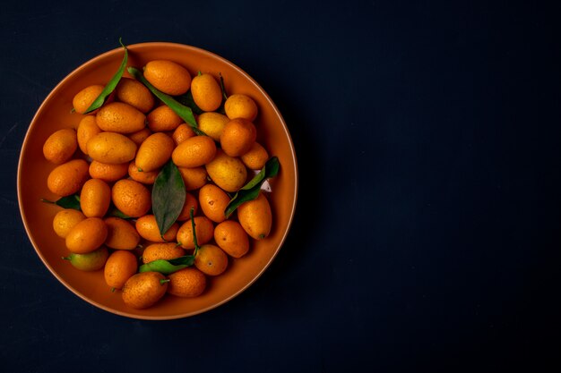 Vista superior de kumquats maduros frescos en un plato sobre superficie negra con espacio de copia